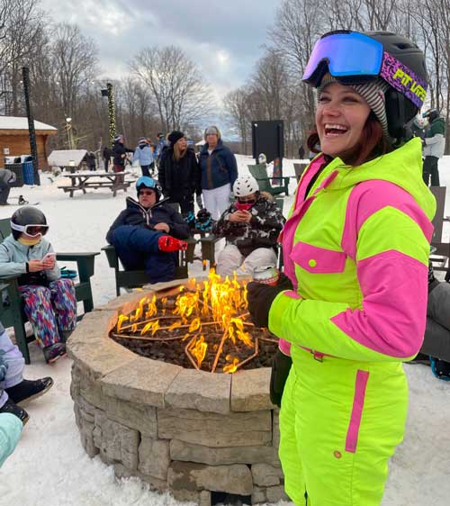 Ashley dressed in bright colored snowsuit gathers with others around a stone firepit