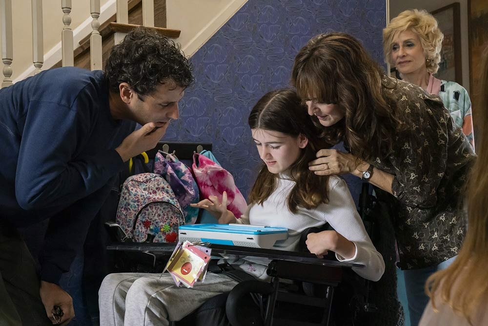 movie scene of mother and father gathered around a girls in a wheel chair looking at a communications device