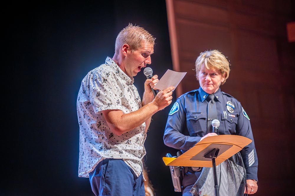 Man reading from a paper on stage with female police officer listening,