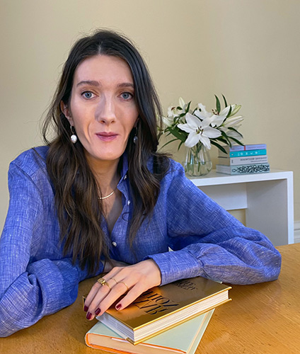 Woman with long hair and a fashionable blue blouse sits at table resting her arm on books on the table