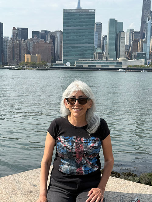 Woman with long gray hair wearing dark sunglasses and a smile sits on a concrete wall with the background of New York city skyline of United Nations and other buildings