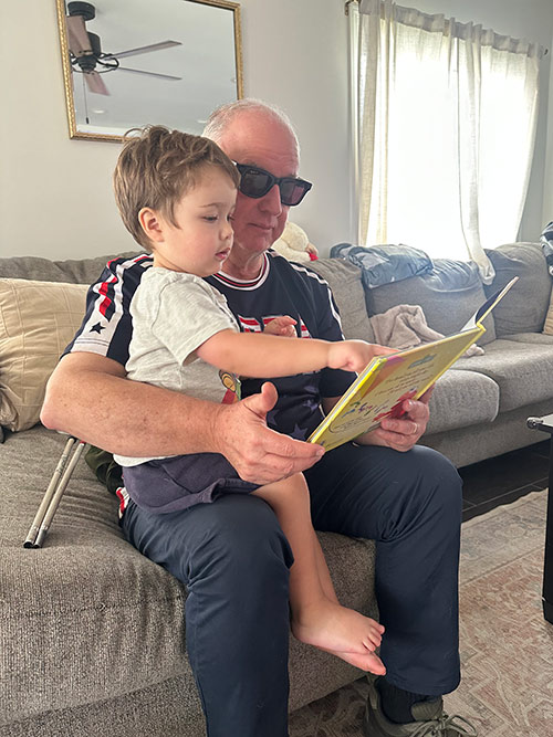 Older gentleman wearing dark sunglasses reads children's book to young boy who is sitting on man's knee on a cozy living room sofa