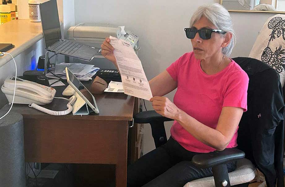 Woman with long gray hair pulled back wearing dark sunglasses sits at a home office desk and looks at a paper utility bill