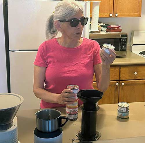 Woman with gray hair pulled back and wearing dark sunglasses stands at kitchen counter looking in the direction if of a food can
