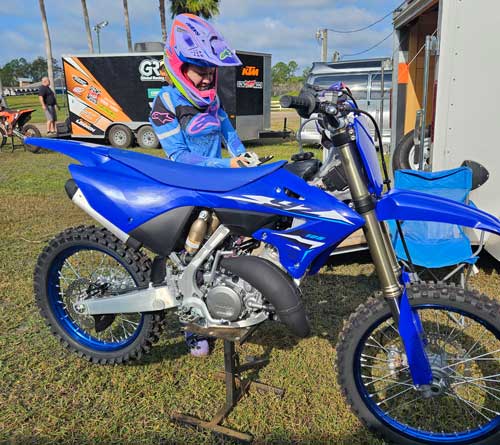 young girl in helmut and dirt bike gear stands on the far side of a bright blue motocross motorcycle
