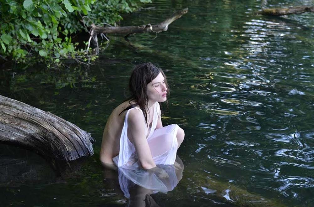 Woman with long hair draped in a white wrapping her arms around her knees while sitting in a gently flowing creek