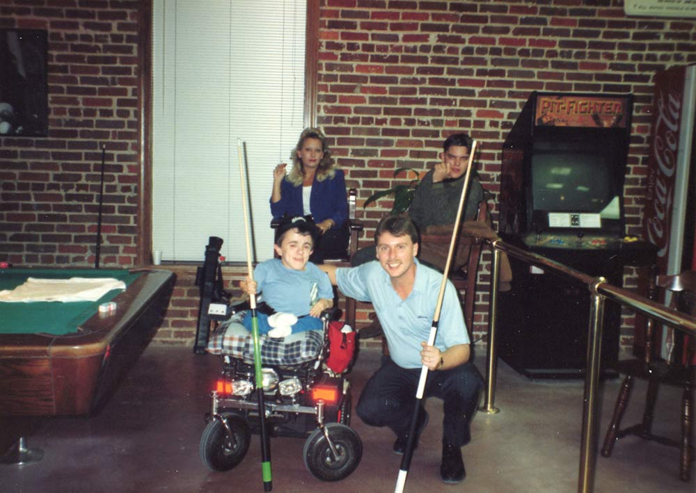 vintage image of young man with diminutive stature smiles and holds a pool stick while sitting in a wheel chair with men and a woman next to a pool table
