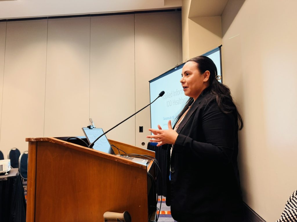 Woman with long hair and dark suit speaks at a podium with a screen to the side