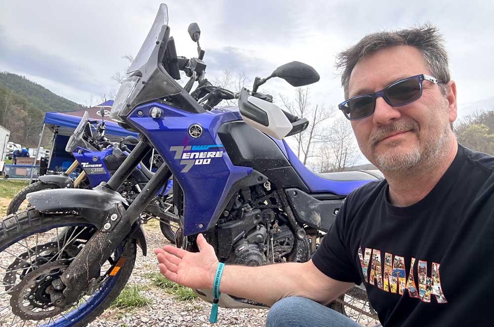 man wearing yamaha t-shirt and sunglasses gestures toward a blue motorcycle in a rural hilly area
