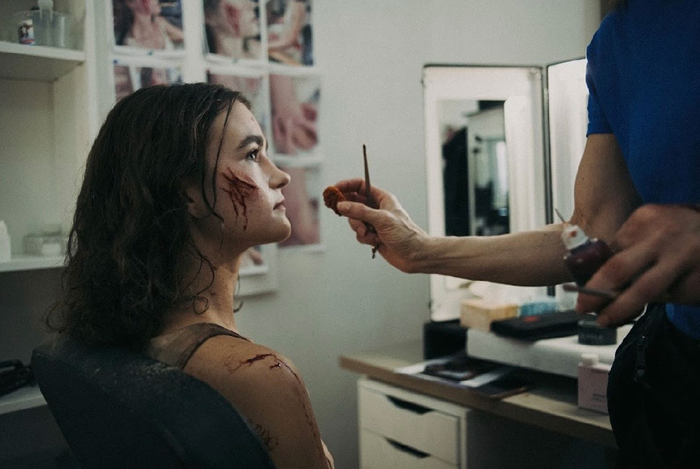 Young woman with long curly hair sits in a chair looking at a make up artist build scars on her face with blood coming from them