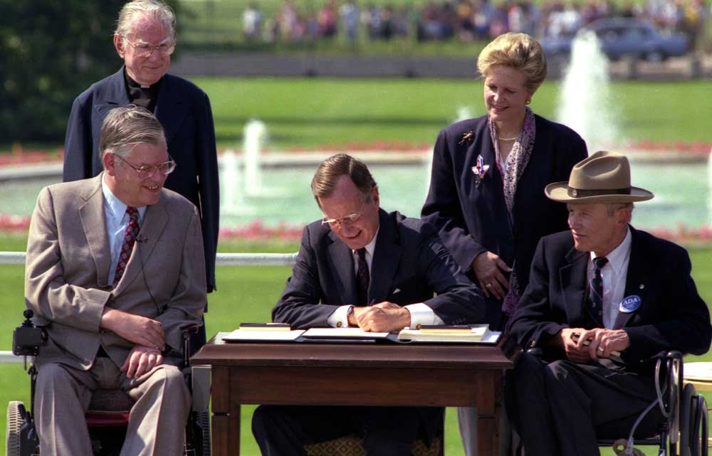 two men in wheel chairs on either side of a man signing a booklet document with a reverend and a woman look on with grass and water fountains in the background