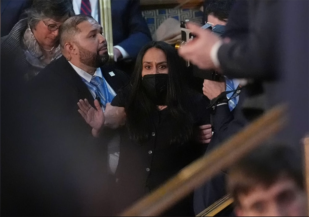 A woman of color with dark hair and a dark face mask is surrounded by people while looking ahead with furrowed brow while a larger man of color with a close shaved beard grabs her arm